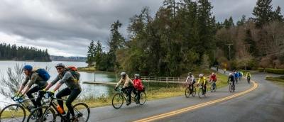 A group of riders cruises along a road next to an inlet that leads to Puget Sound. Docks on the water and evergreen trees define the background.