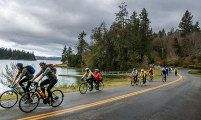 A group of riders cruises along a road next to an inlet that leads to Puget Sound. Docks on the water and evergreen trees define the background.